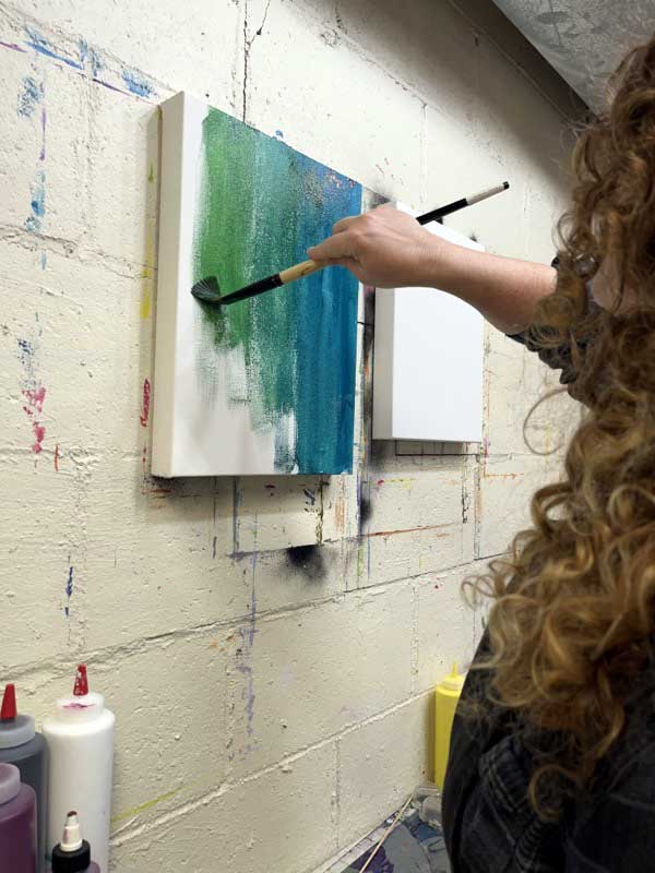 Artist, Jaime Haney, painting the background colors of blue and blue-green on a canvas.